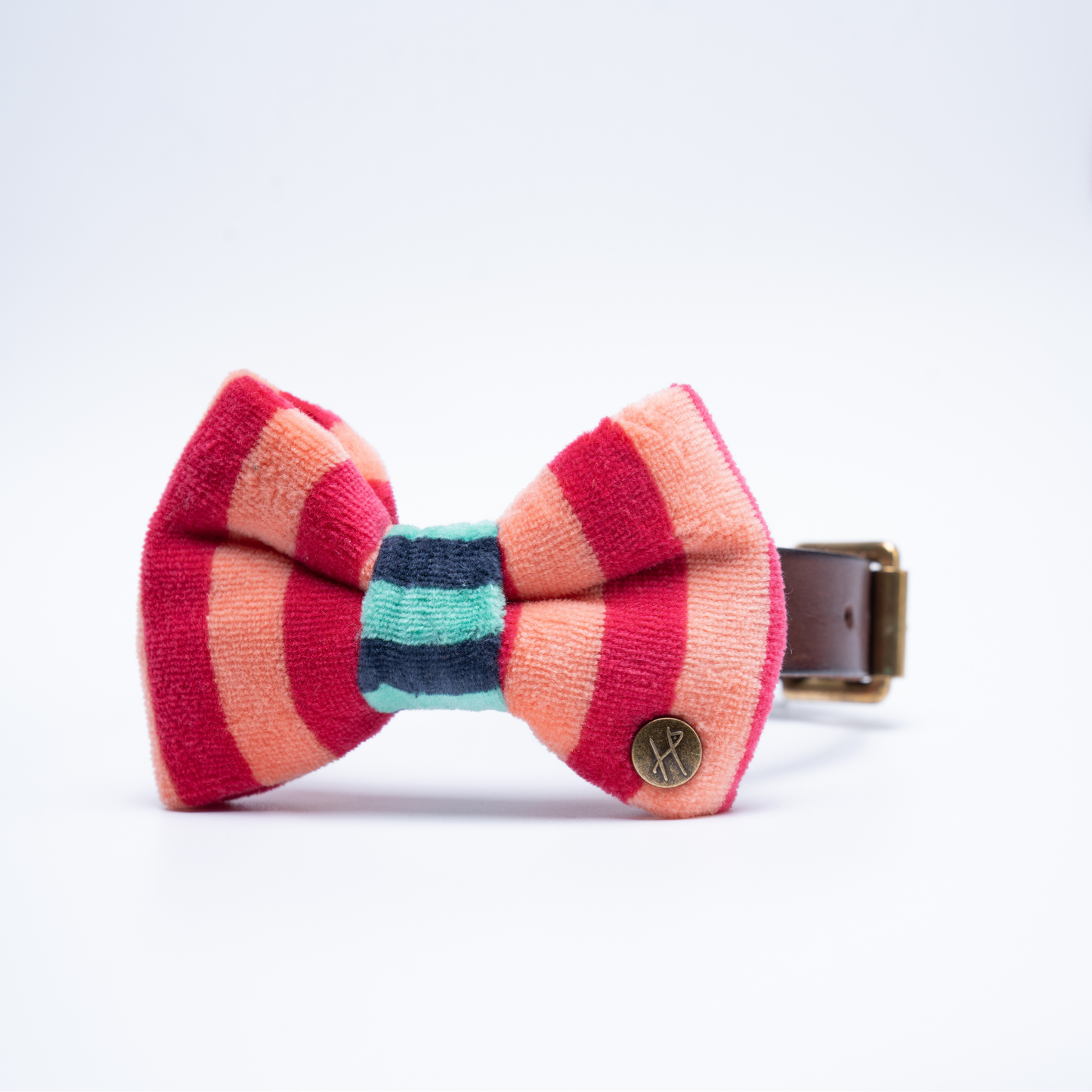A striped bow tie made of textured fabric with alternating pink, coral, and teal stripes, featuring a small round metal logo badge on the right. By HIGGS